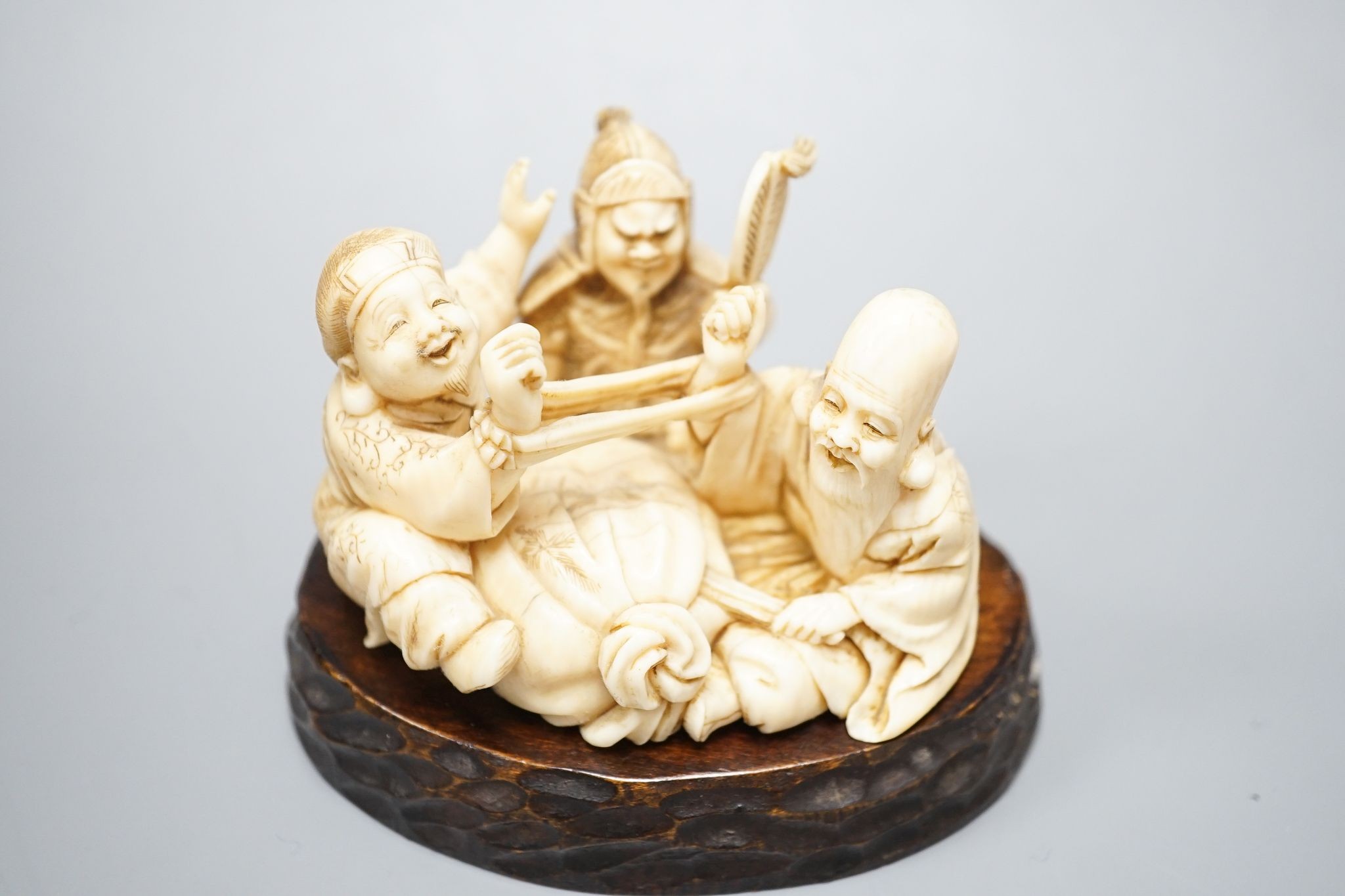 A Japanese ivory okimono of the gods Daikoku, Bishamonten and Fukurokuju, Meiji period, signed to a red lacquer tablet, 8cm wide, wood stand